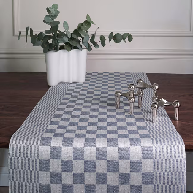 Schackrutan table runner in denim blue on a dark brown dining table in front, with a white vase that has green leaf sticks in it, from Klassbol&#39;s linen weave