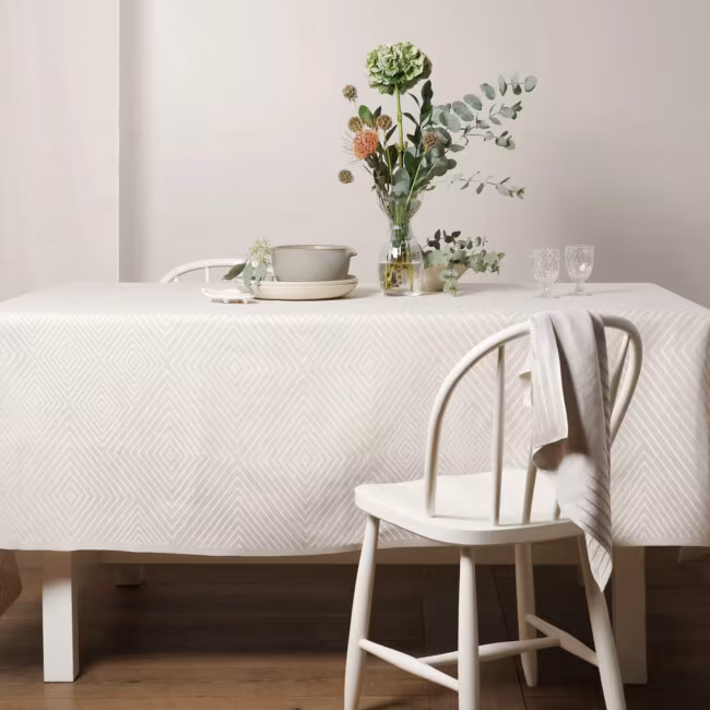 Gåsöga Tischdecke in der Farbe Weiß, auf einem Tisch ausgelegt. Herr ask hängt an einem Stuhl.